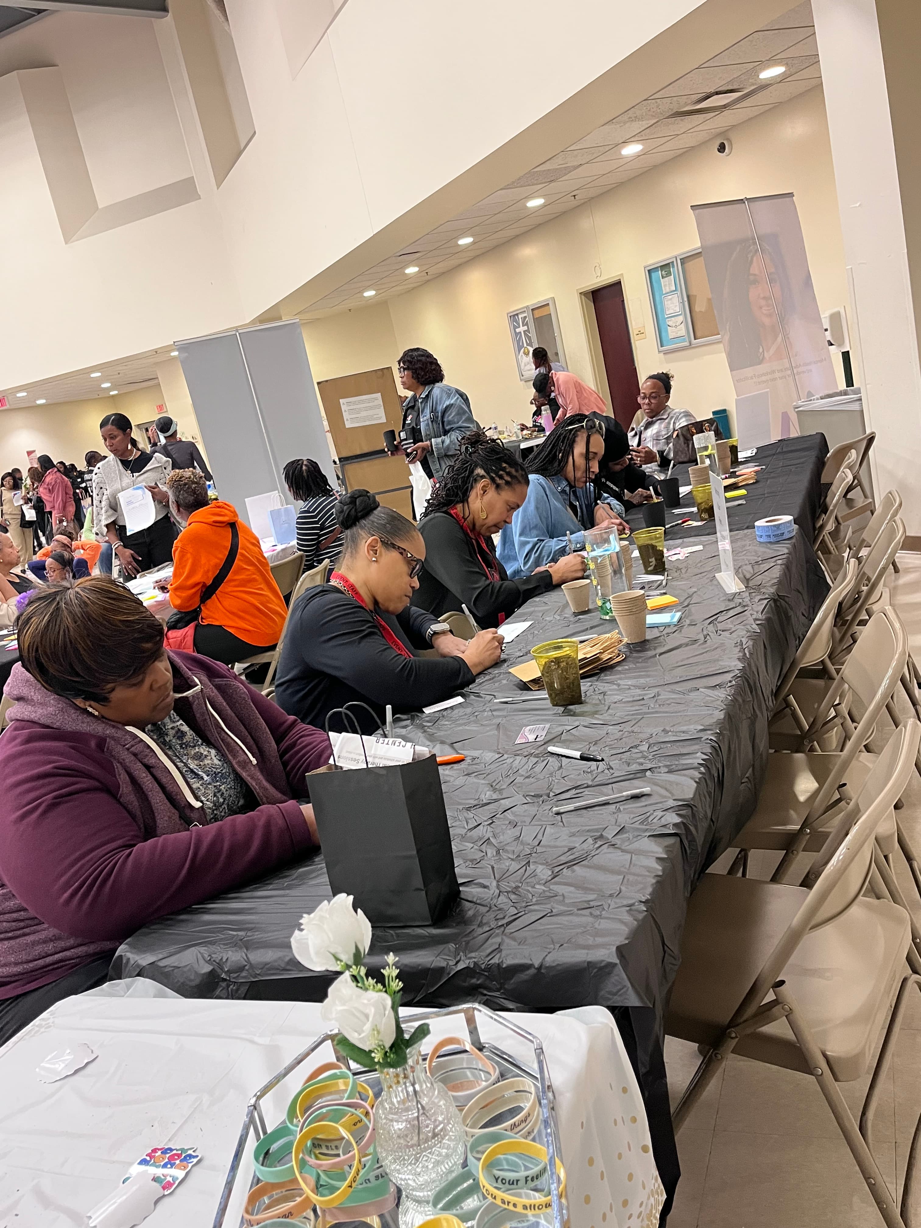 Diverse women sit at a long table with black tablecloth, engaged in a community workshop.