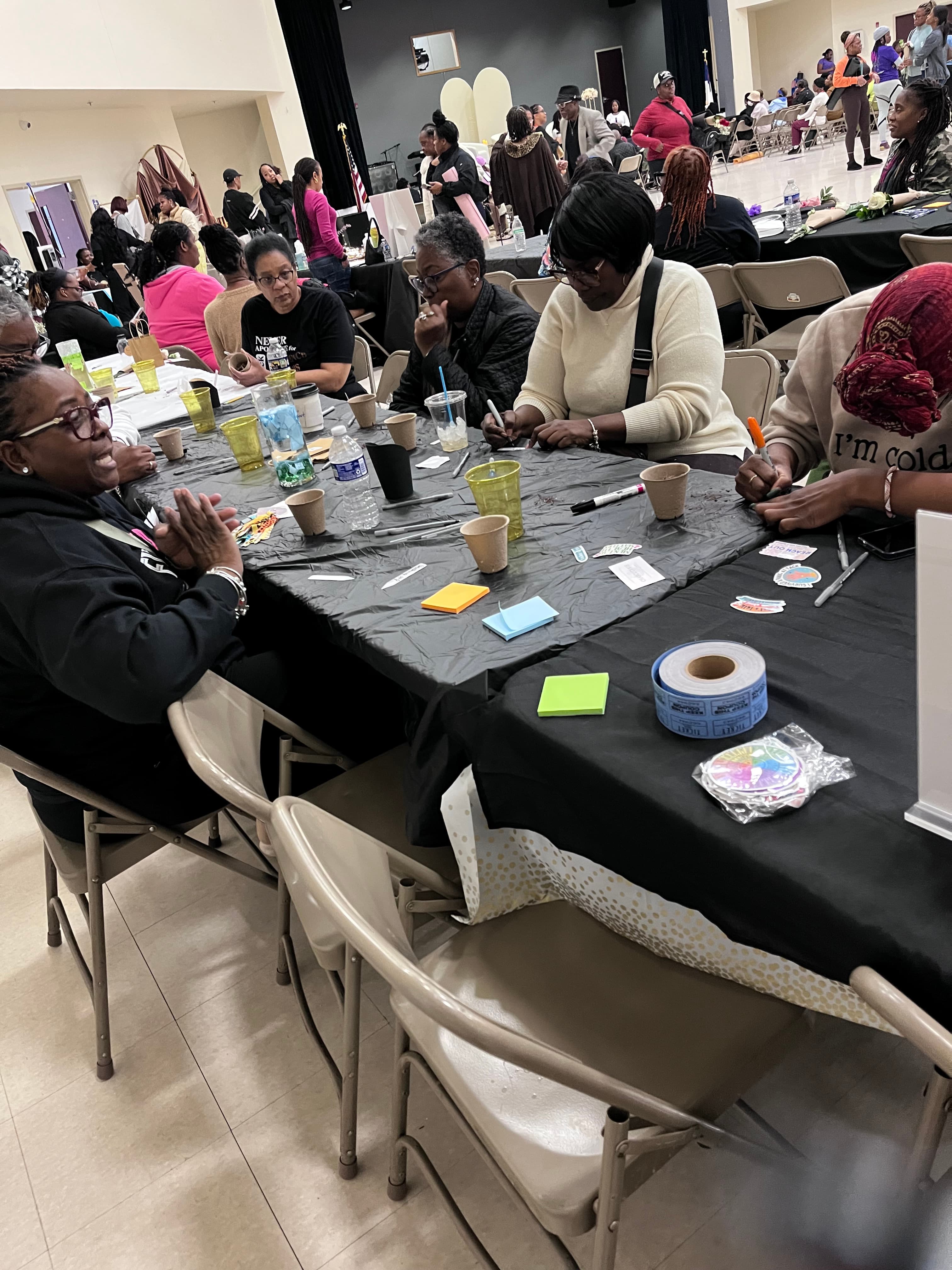 Diverse individuals at black-clothed tables using markers and sticky notes for a collaborative group activity.