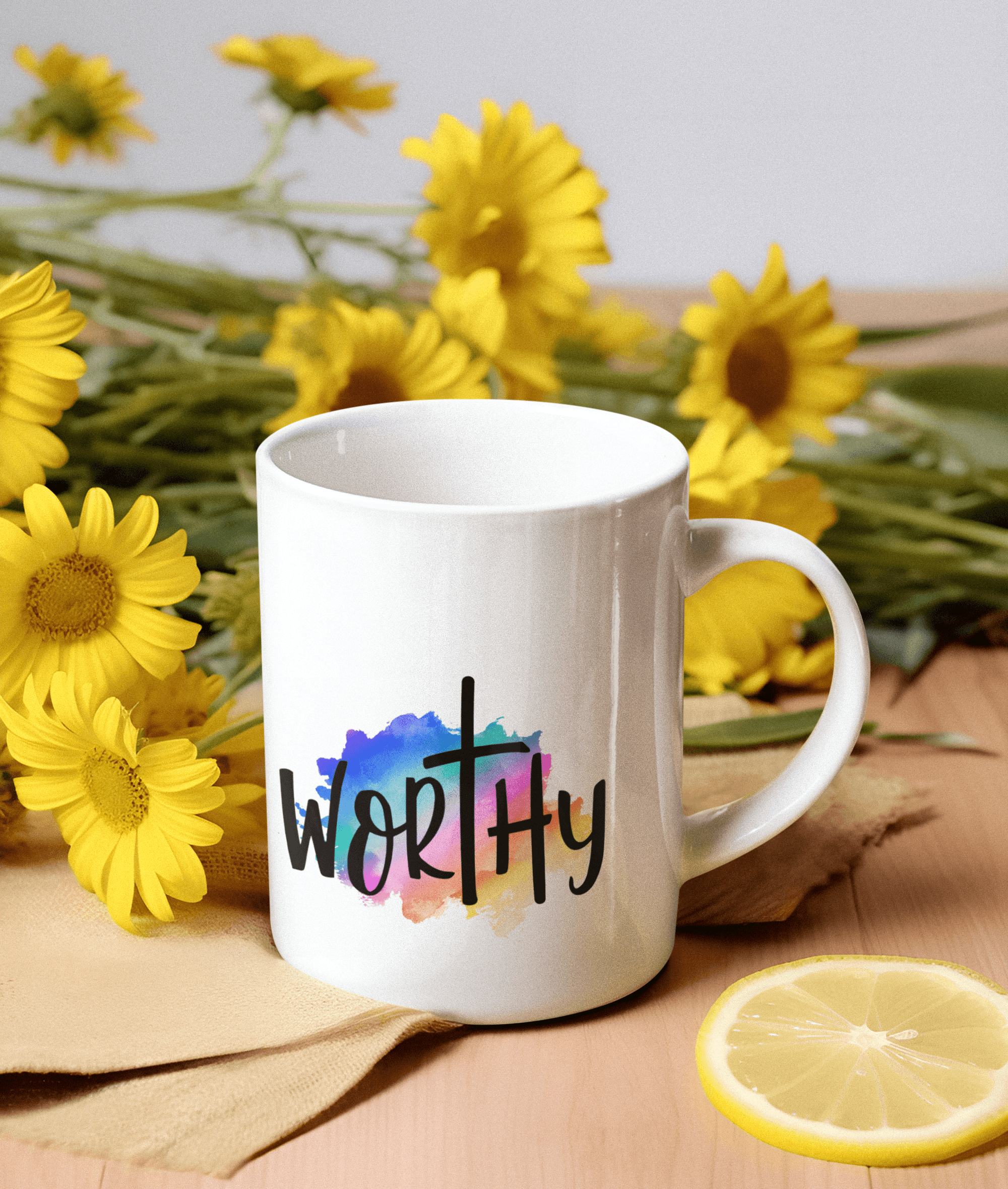White mug with 'WORTHY' and a cross over watercolor, surrounded by vibrant yellow daisies.