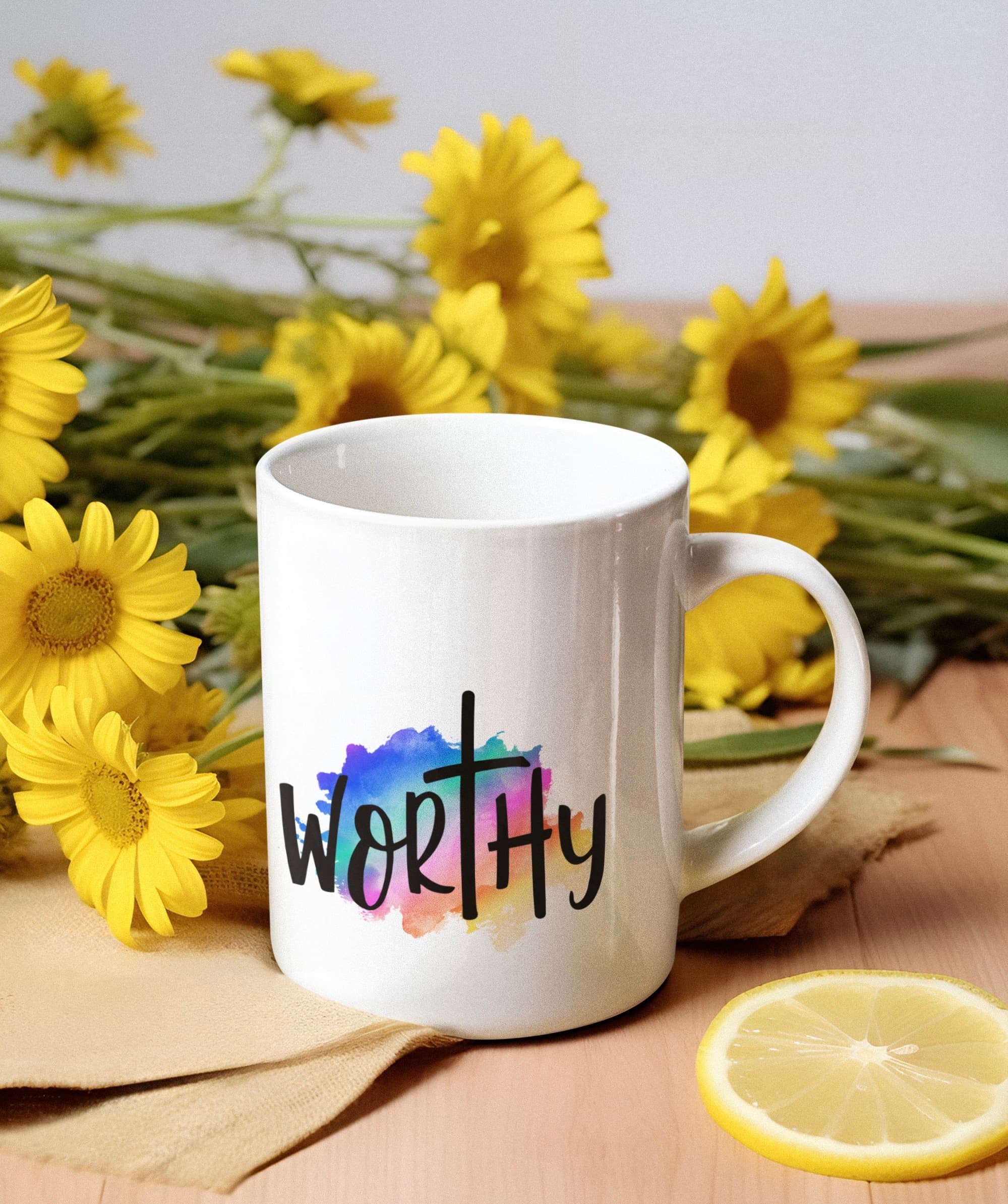 White mug with 'WORTHY' and cross over watercolor splash, surrounded by yellow daisies and lemon.