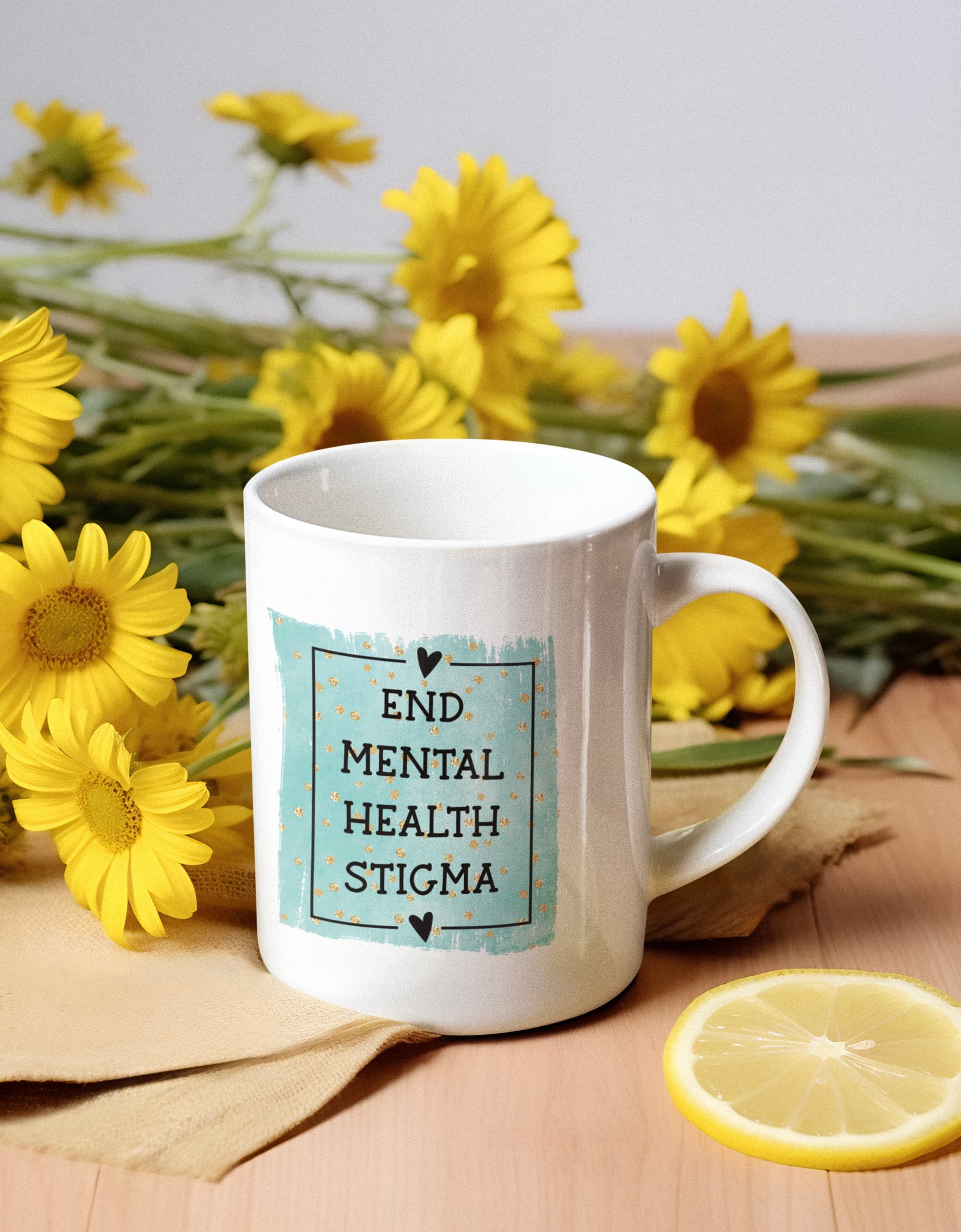 White mug reading "END MENTAL HEALTH STIGMA" surrounded by yellow flowers and a lemon slice.