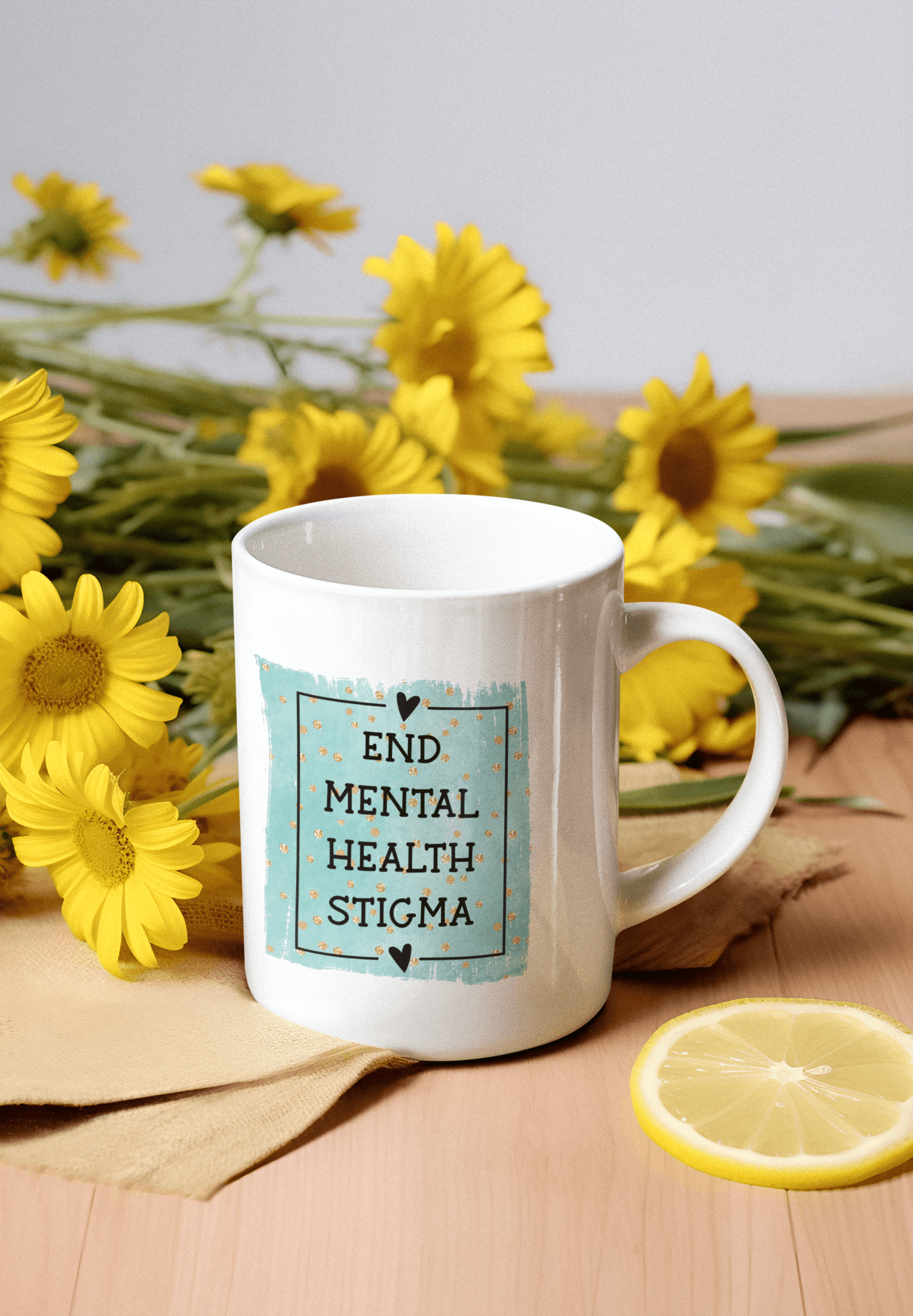 White mug reading "END MENTAL HEALTH STIGMA" surrounded by yellow flowers and a lemon slice.